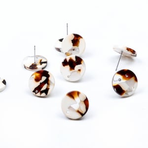 Könnte beinhalten: Eine Sammlung runder Ohrstecker mit einem marmorierten Muster in Braun- und Cremetönen. Die Ohrringe haben silberne Stifte und sind auf einer weißen Oberfläche verstreut. Die Ohrringe sind ein Modeaccessoire.