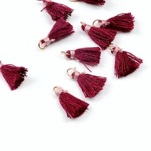 May include: A group of burgundy tassel charms with gold rings. The tassels are made of soft yarn and have a pink accent at the top.