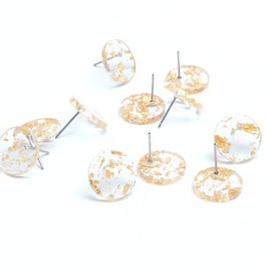 Pode incluir: Um conjunto de dez brincos de flocos de ouro. Cada brinco é um disco redondo de resina transparente com flocos de ouro embutidos. Os brincos são presos a postes prateados.