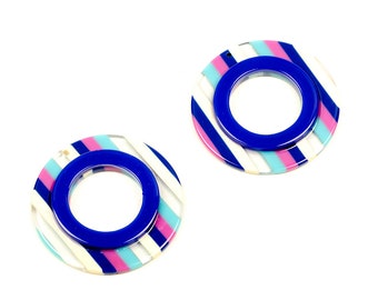 May include: A pair of colorful striped earrings with a blue circle in the center. The earrings are made of acrylic and have a white, pink, and blue striped pattern.
