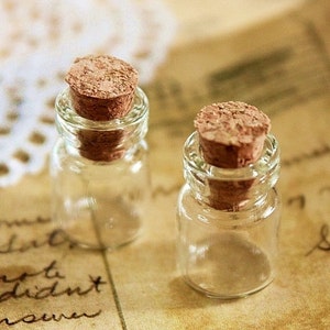 Puede incluir: Dos pequeñas botellas de vidrio transparente con tapones de corcho. Las botellas están sobre un trozo de papel con escritura.