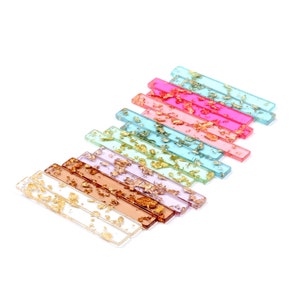 Op de afbeelding: Een set van 12 kleurrijke rechthoekige haarclips van hars met gouden bladeren. De clips zijn gerangschikt in een regenboogpatroon, met kleuren die variëren van lichtblauw tot roze.