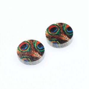 May include: Two round wooden earrings with a peacock feather design. The earrings are brown with green, blue, and red accents.