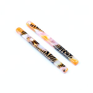 Könnte beinhalten: Zwei lange, dünne Haarsticks mit einem schwarzen, weißen und orangefarbenen Marmormuster.