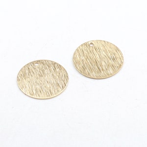 Puede incluir: Dos colgantes redondos de metal dorado con un acabado cepillado texturizado. Cada colgante tiene un pequeño agujero en la parte superior para ensartar. Los colgantes se muestran sobre un fondo blanco. Adecuado para la fabricación de joyas.