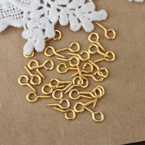May include: A close-up of a pile of gold-colored screw-in eye hooks. The hooks are scattered on a brown surface.