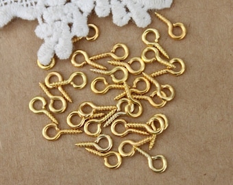 May include: A close-up of a pile of gold-colored screw-in eye hooks. The hooks are scattered on a brown surface.