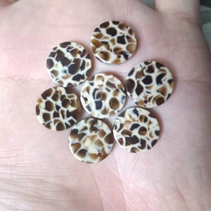 Puede incluir: Siete botones decorativos redondos, planos, con estampado marrón y blanco, con un agujero en el centro.