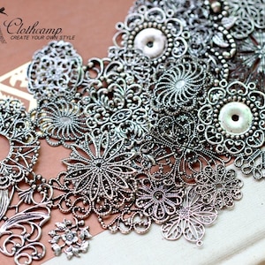 May include: A collection of silver metal filigree charms in various shapes and sizes, including circles, flowers, and swirls. The charms are arranged on a white surface.
