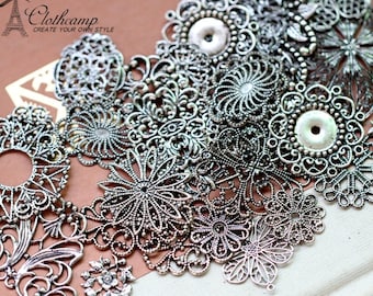 May include: A collection of silver metal filigree charms in various shapes and sizes, including circles, flowers, and swirls. The charms are arranged on a white surface.