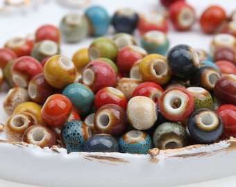 May include: A collection of colorful ceramic beads in various shades of blue, green, red, yellow, and brown. The beads are round and have a smooth, glossy finish. They are arranged in a bowl on a white surface.