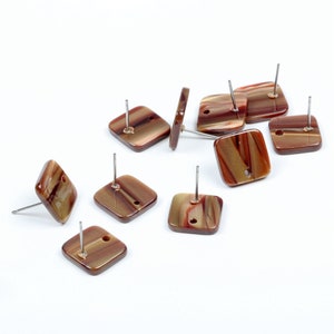 Puede incluir: Un conjunto de diez pendientes cuadrados con patrones marmoleados en marrón, rojo y dorado. Cada pendiente tiene una poste de plata.