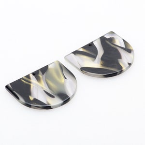 Puede incluir: Dos colgantes de resina de medio círculo marmoleados en negro, blanco y dorado con un agujero en la parte superior para colgar.