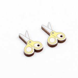 Può includere: Due bottoni di legno gialli a forma di forbici. Le forbici hanno lame bianche e un foro al centro di ogni bottone.