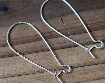 May include: Two silver earring hooks, also known as ear wires, are shown on a wooden surface. The hooks are made of a thin, flexible metal and have a loop at the top for attaching earrings.