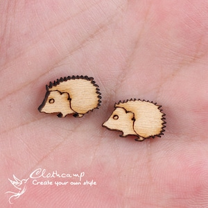 Può includere: Due piccoli ritagli di legno di ricci, ognuno con un design dettagliato e un colore marrone chiaro. I ricci sono rivolti in direzioni diverse e si trovano su una superficie rosa chiaro.