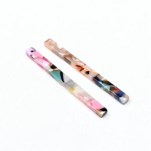 Könnte beinhalten: Zwei rechteckige, farbenfrohe Acrylperlen mit einem Loch an einem Ende. Die Perlen haben ein marmoriertes Muster in Rosa-, Blau-, Grün- und Beigetönen. Ideal für die Schmuckherstellung und das Basteln.