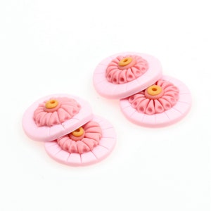 May include: Four pink and yellow floral buttons with a raised design. The buttons are round and have a raised center with a yellow circle in the middle. The buttons are stacked on top of each other.
