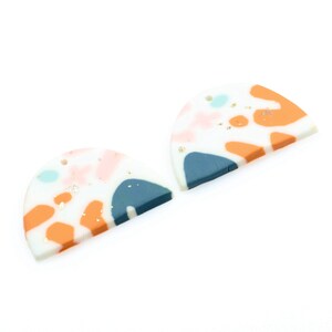 May include: Two half-circle shaped polymer clay earrings with a white base and abstract patterns in shades of orange, pink, blue, and gold glitter.