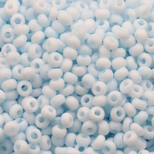 May include: A close-up view of a large quantity of small, light blue glass seed beads. The beads are round and have a hole in the centre.