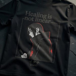 May include: Black t-shirt with an anime character graphic in red and white, holding a weapon. The text "Healing is not linear" is above the graphic, with Japanese text below. The shirt is made of a soft material.
