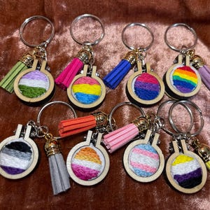 May include: A collection of keychains featuring small, round embroidery hoops with various pride flag designs. Each keychain has a wooden hoop, a metal ring, and a colorful tassel. The flags include rainbow, bisexual, and transgender pride flags.