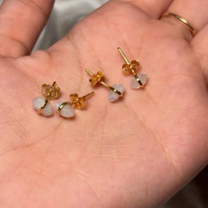 May include: Four pairs of gold-colored stud earrings with small, irregular, light-colored stones. The earrings are displayed on a person's open palm, with a gold ring visible on a finger. The earrings have a simple, elegant design.