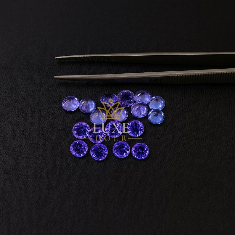 May include: A collection of round, faceted, violet-blue gemstones arranged on a black surface. A pair of silver tweezers is positioned above the gemstones. The gemstones vary in color intensity, with some appearing lighter than others. The image includes the text "LUXE TOUR".