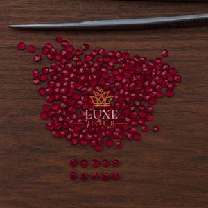 Può includere: Una collezione di piccole gemme rosse scure sfaccettate sparse su una superficie di legno. Le gemme sono disposte in un mucchio sparso con alcune pietre in una fila ordinata. Il testo "LUXE HOUR" è visibile.