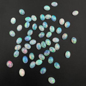 May include: A collection of oval-shaped opals with iridescent colors of blue, green, and pink. The gemstones are scattered on a dark surface, showcasing their polished, smooth surfaces. These opals are ideal for jewelry making.
