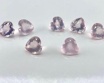 May include: Seven heart-shaped gemstones in a soft pink hue are arranged on a white surface. Each gem has a faceted cut, catching the light and creating a sparkling effect. The gems are likely for jewelry making or crafting.