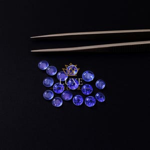 May include: A collection of round, blue-violet gemstones are scattered on a black surface. A pair of tweezers is positioned above the gemstones. The gemstones vary in shade, with some appearing more translucent than others. The Luxe Store logo is visible.