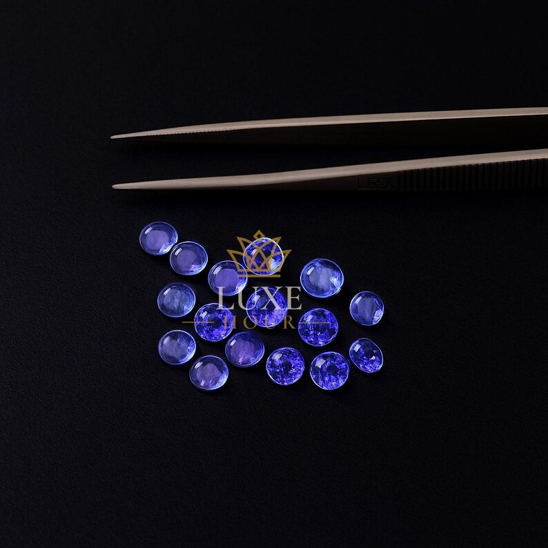 May include: A collection of round, blue-violet gemstones are scattered on a black surface. A pair of tweezers is positioned above the gemstones. The gemstones vary in shade, with some appearing more translucent than others. The Luxe Store logo is visible.