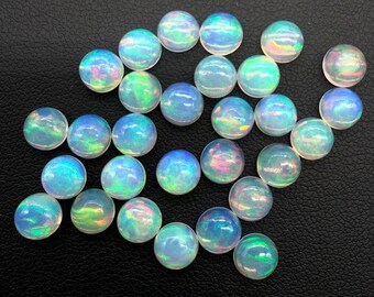 Può includere: Una collezione di pietre preziose di opale rotonde e iridescenti. Gli opali mostrano una gamma di colori, tra cui blu, verde e rosa, con un effetto cangiante. Le pietre preziose sono sparse su una superficie scura.