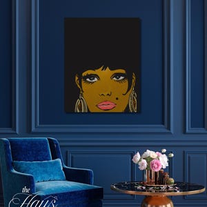 以下が含まれることがあります： 黒い髪と金色の肌色の女性のポップアートスタイルの肖像画。このアートワークは、大きな目、ピンクの唇、ぶら下がっているイヤリングが特徴です。背景は黒です。このアートワークは濃い青色の壁に表示されています。