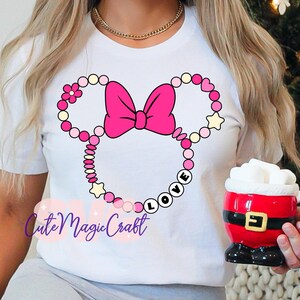 May include: White t-shirt featuring a pink Minnie Mouse design with a bow and the word "LOVE". The design includes pink, yellow, and white beads and stars. A gold necklace is visible. A Santa mug is held in the hand.