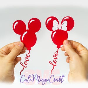 Puede incluir: Dos recortes de globos rojos con forma de Mickey Mouse, uno con un lazo blanco, sostenidos por manos. Cada globo tiene la palabra "love" en escritura cursiva. El texto "Cute Magic Craft" est&aacute; en la parte inferior en morado.