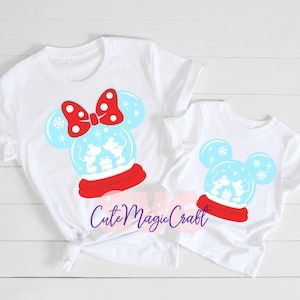 May include: Two white t-shirts with a snow globe design featuring Mickey Mouse silhouettes. The snow globe is light blue with snowflakes, a red base, and a red bow. The text "Cute Magic Craft" is in a cursive font.