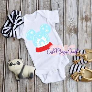 May include: White baby onesie with a Mickey Mouse snow globe design, red scarf, and blue snowflakes. Includes a black and white striped headband, a panda figurine, and gold baby shoes with black and white stripes.
