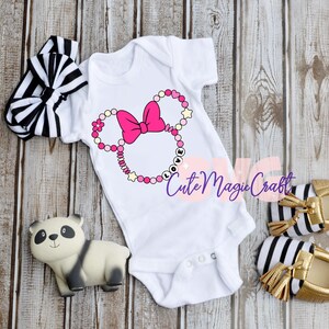 May include: White baby onesie with a pink Minnie Mouse design, a pink bow, and the word "LOVE". A black and white striped headband and gold-trimmed shoes with black and white stripes are also in the image.