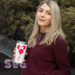 Puede incluir: Un vaso blanco con un dise&ntilde;o de globo de Minnie Mouse rojo, un lazo y la palabra "love" en cursiva. El vaso lo sostiene una persona que lleva un su&eacute;ter burdeos. El fondo es una vista borrosa de follaje verde.