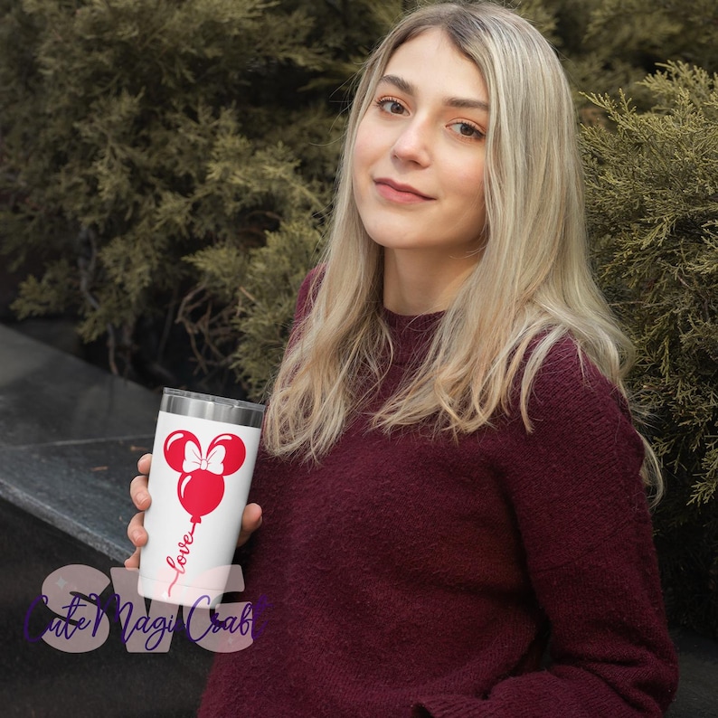 Puede incluir: Un vaso blanco con un dise&ntilde;o de globo de Minnie Mouse rojo, un lazo y la palabra "love" en cursiva. El vaso lo sostiene una persona que lleva un su&eacute;ter burdeos. El fondo es una vista borrosa de follaje verde.