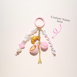 Könnte beinhalten: Ein rosa Schlüsselanhänger mit Charms wie Croissant, Kaffeetasse, Macaron und Eiffelturm. Er hat Perlen, Herzen und den Namen "CHLOE". Der Text "Custom Name here" ist ebenfalls sichtbar. Ein charmantes Accessoire.