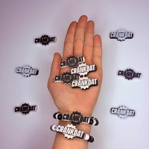 May include: A collection of black and white gear-shaped charms and two bracelets featuring the text CRANKDAT. The charms are arranged in a cluster and scattered around, while the bracelets consist of beads and gear-shaped CRANKDAT plaques.