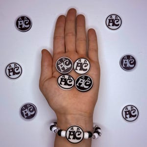 May include: Four circular black and white charms featuring the letters AC with stylized ears, arranged in a square pattern. A matching charm is centered on a beaded bracelet with alternating black and white beads, surrounded by four loose charms.