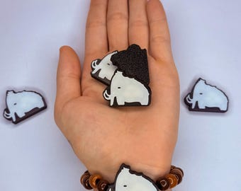 May include: A collection of mammoth-shaped accessories. The image shows several small, dark brown and white mammoth-shaped objects, some held in a hand, and one on a beaded bracelet. The mammoths have white bodies and black outlines.