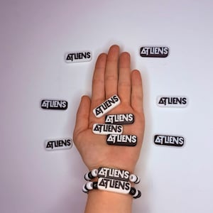May include: A collection of rectangular charms and a beaded bracelet featuring the text ATLIENS. The charms are black with white text or white with black text. The bracelet consists of black and white beads alternating with two rectangular charms.