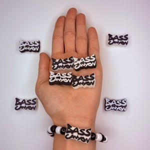 May include: A collection of black and white "BASS CANYON" charms and a bracelet. The charms are in a rectangular shape with the text in a stylized font. The bracelet has black and white beads.