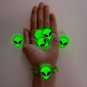 May include: Glow-in-the-dark alien head bracelet and charms. The bracelet is green and yellow with a large alien head charm. Several glowing green alien head charms are scattered around the hand.