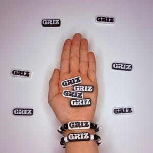 May include: A collection of rectangular charms and two bracelets featuring the text GRIZ in a bold white font on a black background. The charms are scattered around, while the bracelets are worn on a wrist, showcasing the repeating logo design.